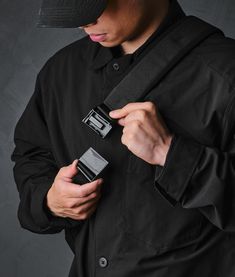 a man wearing a black hat and jacket holding two cell phones in his hands while standing against a gray background