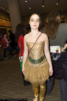 a woman wearing gold pants and a dress with an eye patch on her face is standing in front of a group of people