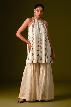 White and gold halter neck organic cotton top embellished with antique gold aari and zardozi work using zari thread, beads and sequins. Paired with zari silk striped sharara pant and brown silk dupatta.
Components: 3
Pattern: Embroidery
Type Of Work: Aari, Zardozi
Neckline: Halter Neck
Sleeve Type: Sleeveless
Fabric: Organic Cotton, Zari Cotton Silk
Color: White, Gold
Other Details: 
Attached inner lining
Length: 
Kurta: 34inches
Sharara pant: 42inches
Model Height: 5ft 9inches, wearing size M
O Festive Sleeveless Cotton Palazzo Set, Traditional Sleeveless Cotton Palazzo Set, Sleeveless Cotton Sharara For Diwali, Bohemian Sleeveless Sharara For Diwali, Bohemian Sleeveless Sharara For Festive Occasions, Festive Gold Cotton Sharara, Festive Bohemian Sleeveless Sharara, Bohemian Sleeveless Sharara For Navratri, Festive Sleeveless Cotton Sharara