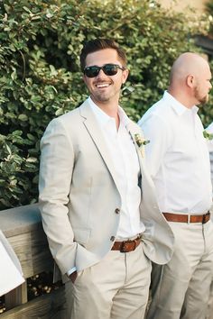 two men in suits and sunglasses standing next to each other on a wooden bench with bushes behind them