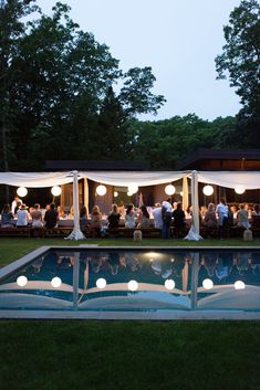 a group of people sitting at tables next to a swimming pool with lights on it