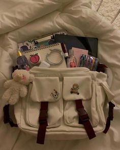 a white purse filled with lots of items on top of a bed next to a teddy bear