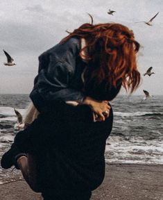 a woman with red hair is standing on the beach and birds are flying around her