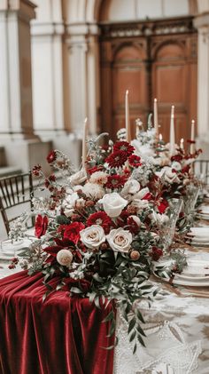 an elegant table setting with red and white flowers, greenery and candles on it