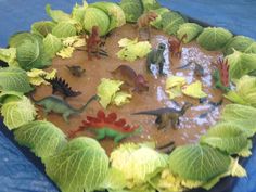 an assortment of toy dinosaurs and plants in a plastic tray on a blue tablecloth