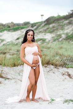 a pregnant woman in a pink dress standing on the beach with grass and sand behind her