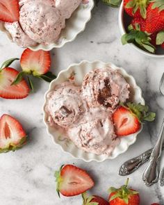 two bowls filled with ice cream next to strawberries on a marble counter top and spoons