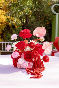 red and pink flowers are in a vase on a white tablecloth with an orange chair behind it