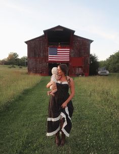 a woman holding a baby in front of a red barn with an american flag on it