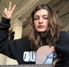 a woman with long hair is holding a guitar and making the peace sign in front of her