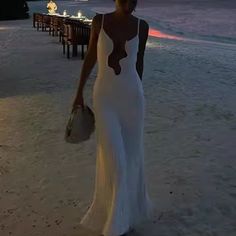 a woman in a white dress is walking on the beach at sunset with her purse