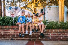 three children sitting on a brick wall with mickey mouse balloons in front of their heads