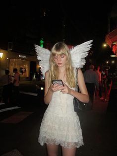 a woman dressed as an angel checks her cell phone while standing in the street at night