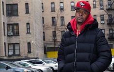 a man in a black jacket and red hat standing on the street next to parked cars