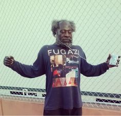 a man with his hands out standing in front of a chain link fence holding a can of fugaz