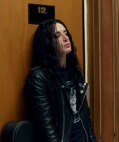 a man with long black hair sitting in front of a door and looking off to the side