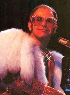 a man with glasses and a fur vest on holding a microphone in front of him