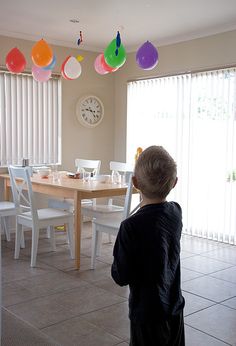 a little boy standing in front of a dining room table with chairs and balloons hanging from the ceiling