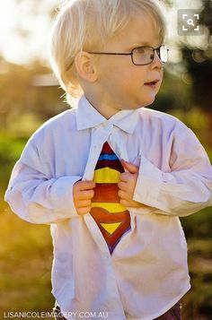 a little boy in glasses and a superman shirt is holding his tie up to his chest