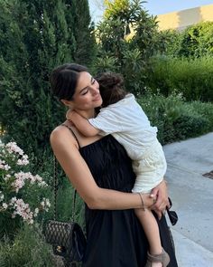 a woman holding a child in her arms while standing on a sidewalk next to flowers