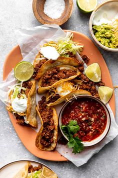 tacos and guacamole are served on an orange plate with lime wedges