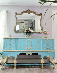 an ornate blue and gold dresser with flowers on top, in front of a mirror