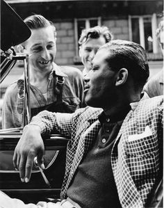 black and white photograph of four men sitting in a car talking to one another while the man is holding a microphone