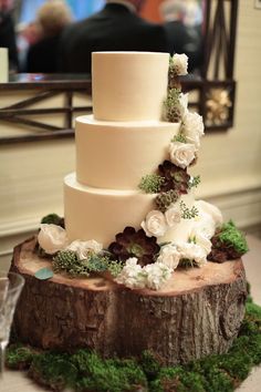 a three tiered wedding cake with white flowers and greenery on a tree stump