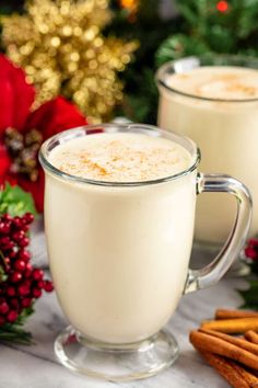 two mugs of eggnog with cinnamon sticks and christmas decorations in the background