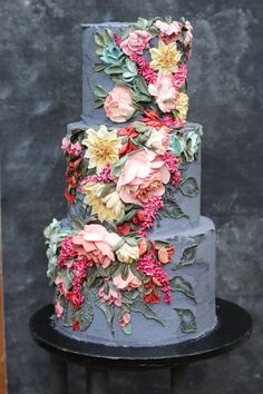 a three tiered cake decorated with flowers on a black stand in front of a gray wall