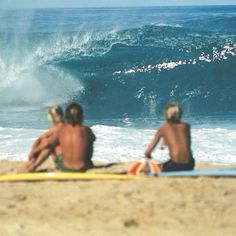 three people sitting on the beach watching a wave