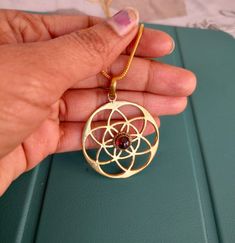 a hand holding a gold pendant with a flower on it's end and a red stone in the center