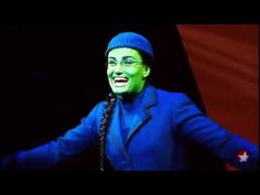 a man with green paint on his face wearing a blue suit and hat, standing in front of a black background