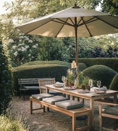 an outdoor dining table with chairs and umbrella in the middle of a garden area surrounded by hedges