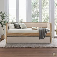 a daybed with pillows and blankets on it in front of two windows, next to a potted plant