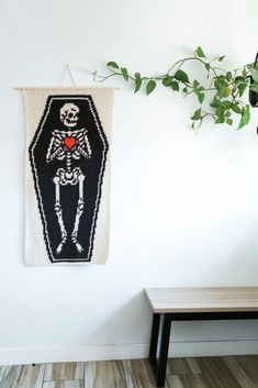 a cross - stitch skeleton hanging on the wall next to a table with a potted plant