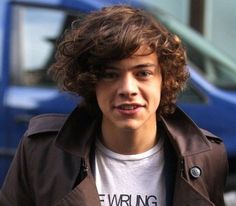 a young man with curly hair wearing a brown jacket and white t - shirt standing in front of a blue truck