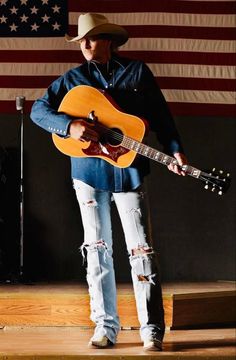 a man with a cowboy hat holding an acoustic guitar in front of an american flag