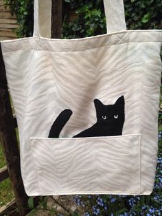 a black cat is sitting in the pocket of a white tote bag with blue flowers behind it