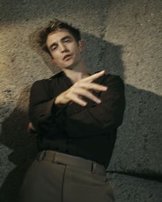 a young man leaning against a wall with his arms stretched out to the side, wearing a black shirt and khaki pants