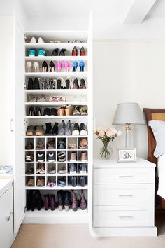 a white closet filled with lots of shoes next to a nightstand and lamp on top of a bed