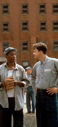 three men standing next to each other in front of a brick building and one is holding his hands together