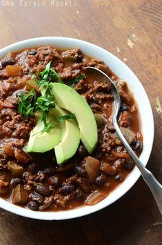 a white bowl filled with chili and avocado on top of a wooden table