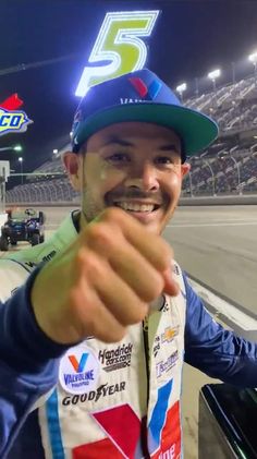 a man pointing at the camera in front of a race track with other cars behind him