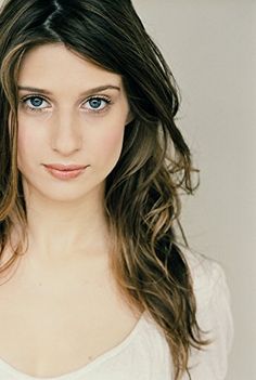 a woman with long brown hair and blue eyes posing for a photo in front of a white wall