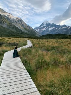 a person sitting on a wooden walkway in the middle of a grassy field with mountains in the background