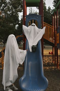 a person in a ghost costume standing next to a slide at a play ground with swings and slides