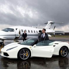 two men standing next to a white sports car in front of an airplane on the tarmac