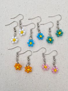 six pairs of flower earrings with dangling hooks on a white cloth covered surface, all in different colors and sizes