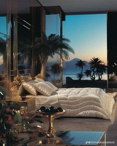 a bedroom with large windows overlooking the ocean and palm trees at sunset or dawn,